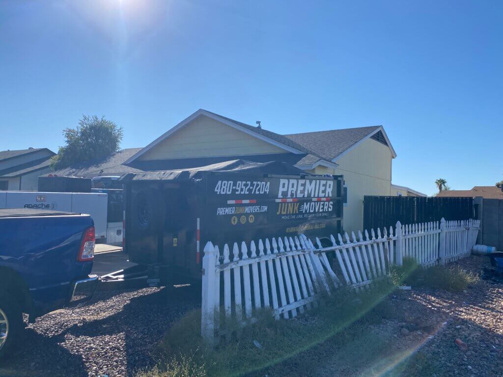 photo of a premier dumpster rental staged in front of a customers home. The trailer is parked next to a white picket fence and sky is sunny and blue.